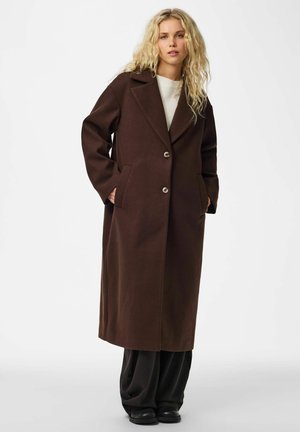 Une femme blonde porte un long manteau marron foncé boutonné par-dessus un haut blanc et un pantalon noir ample, sur un fond de studio uni.