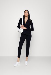 Blazer noir ajusté, pantalon noir moulant et baskets blanches. Le modèle tient un sac bandoulière blanc. Tissu lisse, design minimaliste.