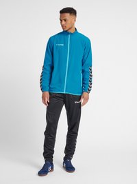 Lichtblauwe sportjas met hoge kraag en zijrits, voorzien van zwarte chevronpatronen op de mouwen; gecombineerd met zwarte sportbroeken.