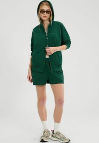 Grüne Zip-Hoodie und passende Shorts aus weichem Stoff. Verfügt über eine Kapuze, Seitentaschen und ein Logo auf der oberen linken Brust.