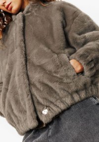 Veste en fausse fourrure grise avec une texture douce et pelucheuse, col montant et poches latérales. Présente un bouton rond en argent comme détail de fermeture.