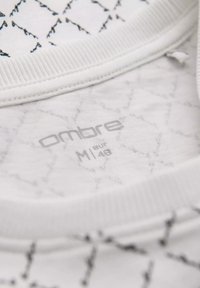 Bílá látka s geometrickým černým vzorem, se žebrovaným výstřihem. Štítek nese nápis "ombre" a velikost "M/eur 48". Měkká textura.