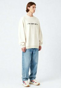 Felpa oversize di colore crema con testo nero "HO PARLATO A CHATGPT DI TE", abbinata a jeans azzurri larghi e sneakers bianche con accenti rossi e blu.