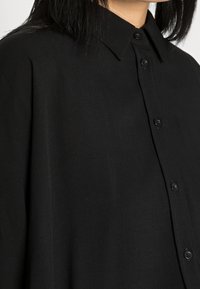 Camicia nera con bottoni, realizzata in tessuto testurizzato, con colletto classico e bottoni frontali. Il design è ampio con una silhouette drappeggiata.