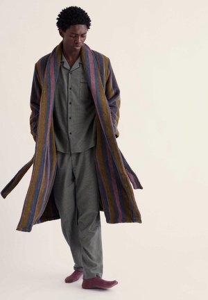 Homme portant un pyjama gris, des chaussettes violettes et une longue robe rayée aux couleurs bleu, rose et moutarde, debout devant un fond uni.