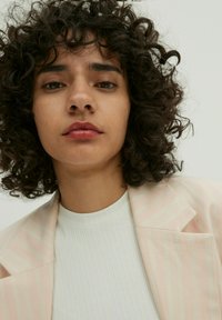 Hellrosa gestreifter Blazer mit einem strukturierten weißen Oberteil darunter. Das Model hat lockige Haare und einen neutralen Hintergrund.