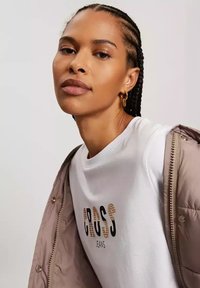 Biała koszulka z grafiką "CROSS JEANS" w czerni i pomarańczy, zestawiona z jasnobrązową puchową kurtką, z zamkiem błyskawicznym i zatrzaskami.