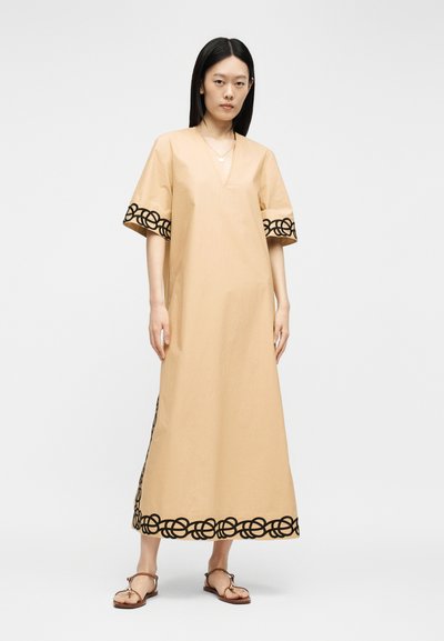 Femme portant une robe beige longueur cheville avec des motifs circulaires noirs brodés sur les manches et l'ourlet, associée à des sandales plates marron.