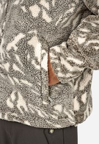 Polarowa kurtka w kolorze szarym i białym z teksturowaną, puszystą powierzchnią; posiada boczną kieszeń i luźny krój.