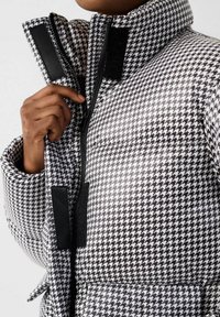 Chaqueta acolchada con un patrón de pata de gallo en blanco y negro, que cuenta con un cuello alto, cremallera frontal y detalles en negro con cierre de gancho y bucle.