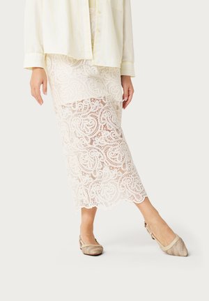 Kobieta w kremowej koronkowej spódnicy midi z falowanym brzegiem, w połączeniu z beżowymi, spiczastymi balerinami na pasku oraz jasnżółtą, zapinaną na guziki koszulą.