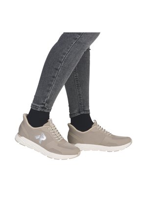 Beige Sportschuhe mit Mesh-Obermaterial, weißer Sohle und Schnürdesign. Merkmale sind ein reflektierendes Logo und strukturierte Akzente an den Seiten.