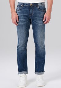 Jean en denim bleu foncé avec une coupe fuselée, texture délavée, design à cinq poches et couture dorée, assortis à des baskets blanches.