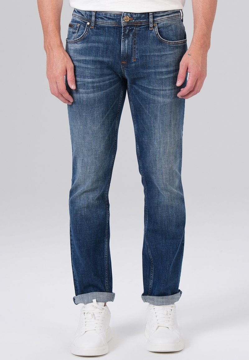 Jean en denim bleu foncé avec une coupe fuselée, texture délavée, design à cinq poches et couture dorée, assortis à des baskets blanches.