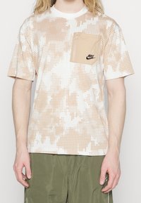 Beige och vit t-shirt med korta ärmar som har ett prickmönster och en ficka med dragkedja på bröstet med en svart logotyp. Mjuk tyg, avslappnad passform.