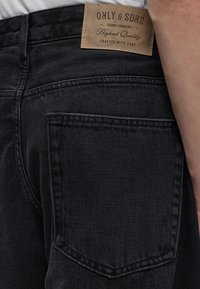 Гръб на черни дънки с кафява кожена лепенка с надпис ""Only & Sons Denim Company Highest Quality Crafted with Care.""