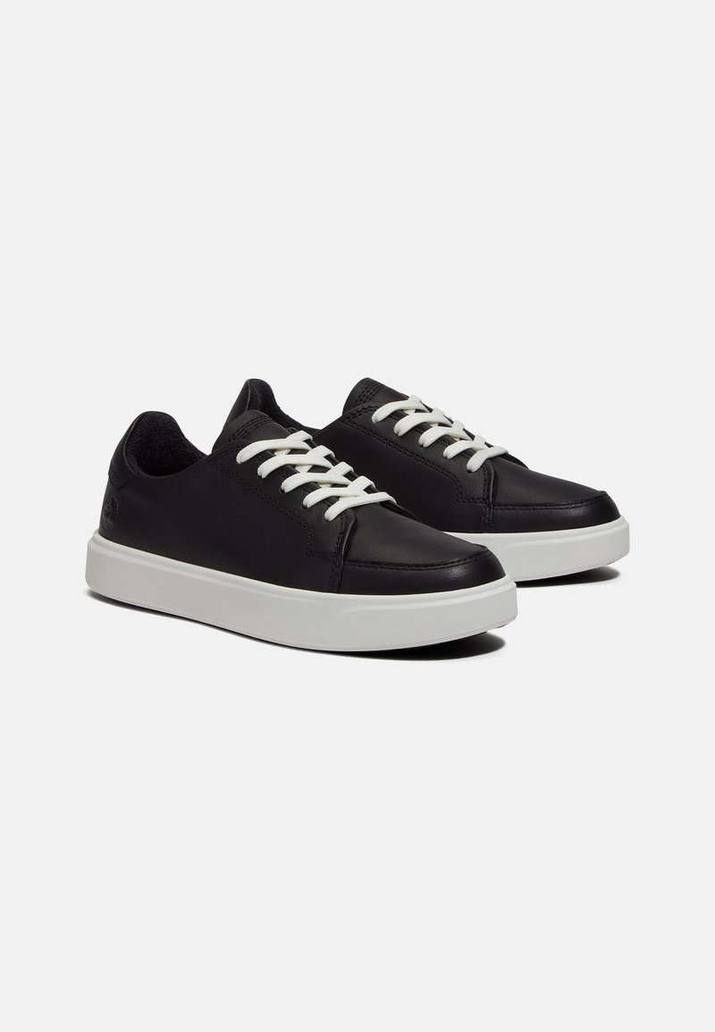 Zapatillas de cuero negro con cordones blancos, punta redonda y suela texturizada. Presentan un logo en el lateral. Diseño simple y de perfil bajo.