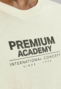 Kreminės spalvos medvilnės marškinėliai su apvaliu iškirpimu; juodas grafikos tekstas, skaitantis "PREMIUM ACADEMY INTERNATIONAL CONCEPT SINCE - 1990."