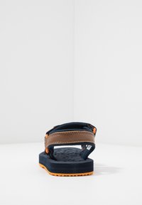 Námořnické žabky s hnědým koženým páskem, texturovanou černou stélkou a oranžovou podrážkou. Otevřený design s nastavitelným zapínáním a polstrovanou oporou.