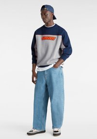 Tröja i marinblått och grått med orange "VANS"-logotyp; kombinerad med lösa ljusblå jeans och svarta-vita skor. Kepa i matchande marinblå färg.