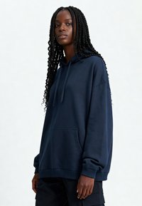 Felpa blu navy con tasca a marsupio, texture morbida, cappuccio con cordino e polsini a coste. Presenta una vestibilità rilassata e oversize.