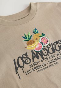 Sudadera de algodón color caqui con un colorido diseño de frutas bordadas (piña, sandía, plátano) y texto impreso en negro.