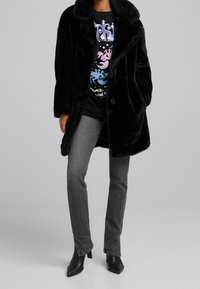 Person wearing a black faux fur coat, graphic black t-shirt, gray jeans, and black heeled ankle boots against a light gray background.