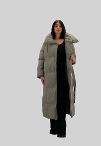 Abrigo largo acolchado en verde apagado, con un cuello alto, textura acolchada y un cinturón en la cintura, llevado sobre una camiseta negra y pantalones de pierna ancha.