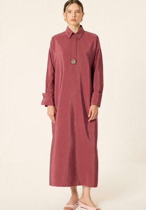 Femme debout, face à l'avant, portant une robe mauve à manches longues, longueur cheville, avec un col, un collier pendentif et des sandales rose clair.