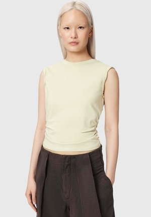 Femme aux cheveux blonds platine et raides portant un haut sans manches jaune pâle et un pantalon ample taille haute foncé sur un fond uni.
