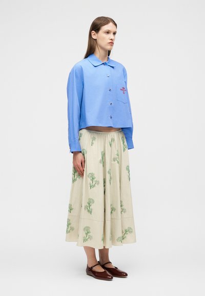 Γυναίκα στέκεται φορώντας ένα ανοιχτό γαλάζιο μακρυμάνικο πουκάμισο με κουμπιά και μια μπεζ μίντι φούστα με πράσινα λουλουδένια σχέδια και καφέ επίπεδα παπούτσια.