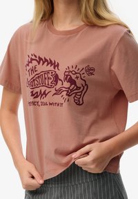Mujer tirando del dobladillo de una camiseta color rosa polvo con texto gráfico burdeos que dice "The Hot Stuff" y una ilustración de serpiente, vestida con pantalones grises de rayas finas.