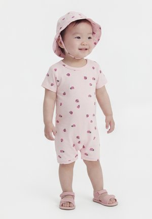 Väikelaps seisab, kannab roosat mütsi, täpikestega kombekat ja sobivaid roosasid sandaale, naeratab ja vaatab veidi paremale.