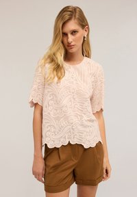 Donna bionda che indossa una blusa leggera ricamata di un rosa chiaro e pantaloni corti con vita alta marroni, su uno sfondo neutro.