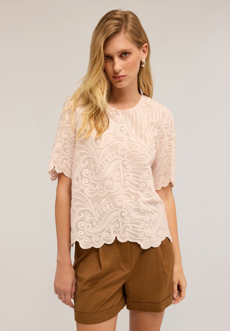 Donna bionda che indossa una blusa leggera ricamata di un rosa chiaro e pantaloni corti con vita alta marroni, su uno sfondo neutro.