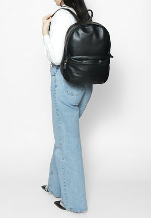Zwarte leren rugzak met een ronde vorm, een ritsvak aan de voorkant en dubbele schouderbanden, gedragen door iemand in een blauwe flared jeans.