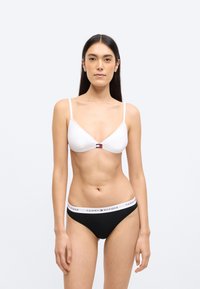 Top de bikini alb în formă de triunghi cu bretele subțiri, având un mic logo Tommy Hilfiger. Partea de jos a bikiniului neagră cu o bandă albă cu logo-ul brandului.