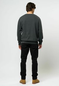 Sötétszürke pulóver, kerek nyakkivágással, bordázott mandzsettákkal és aljjal. Fekete nadrággal és barna bokacsizmával viselve, hátulról látható, világos háttér előtt.