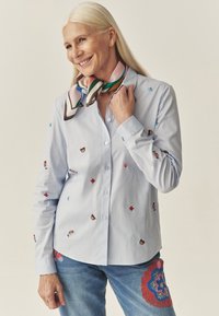 Jasnoniebieska koszula z długim rękawem z kolorowymi haftowanymi motywami; charakteryzuje się gładką tkaniną. Połączona z dżinsami z wyrazistym haftem.