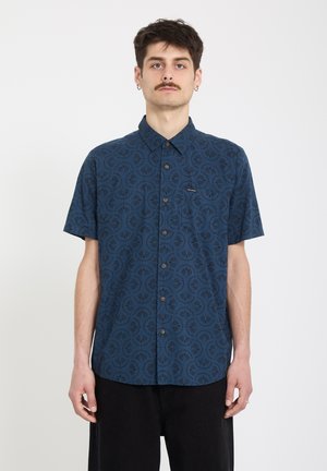 Junger Mann mit kurzen Haaren und Schnurrbart, der ein blau gemustertes, kurzärmeliges Hemd mit Knöpfen und schwarze Hosen trägt, vor einem weißen Hintergrund steht.