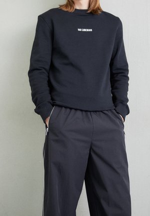 Person trägt schwarzen Langarm-Sweatshirt mit weißem Text und dunkle, lockere Hose mit Reißverschlusstaschen, Hände in den Taschen, steht an einer Wand.