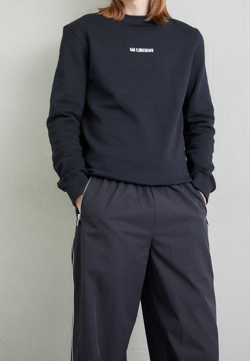 Personne portant un sweat-shirt noir à manches longues avec un texte blanc et un pantalon ample foncé avec des poches zippées, mains dans les poches, debout contre un mur.