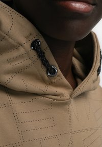 Sudadera con capucha de tela en color marrón claro con costuras negras y patrones de diseño. Presenta un cordón negro y ojales cerca del cuello. Textura suave.