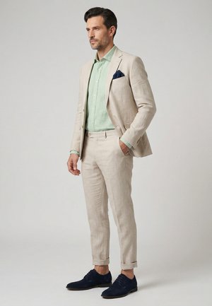 Mann in beigefarbenem Leinenanzug mit mintgrünem Hemd, navy Einstecktuch und navy Herrenschuhen vor hellgrauem Hintergrund.