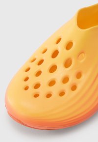 Κίτρινο παπούτσι χωρίς κορδόνια με κυκλικά οπτικά αερισμού. Το λείο υλικό, που μοιάζει με πλαστικό, μεταβαίνει σε πορτοκαλί διαβάθμιση στη σόλα.