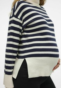 Suéter de rayas en azul marino y blanco con un cuello alto, que presenta aberturas laterales y una textura suave, llevado sobre pantalones negros.