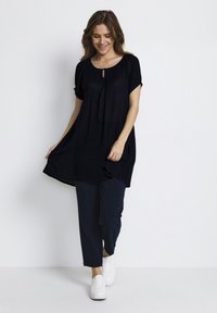Tunique noire à manches courtes, avec des détails froncés au niveau du col, coupe décontractée, associée à un pantalon marine et des baskets blanches. Tissu léger.