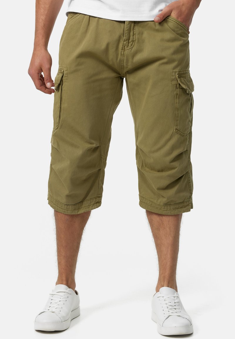 Olivenfarbene Cargo-Shorts aus Baumwoll-Mischgewebe, mit seitlichen Taschen, knielangem Schnitt und gestickten Details. Kombiniert mit weißen Sneakern.
