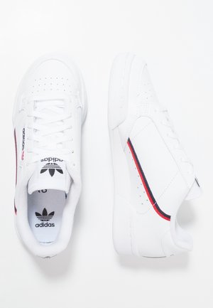 Par de zapatillas blancas Adidas con franjas laterales rojas y azules, puntera perforada y logo negro de Adidas en la lengüeta y la plantilla.
