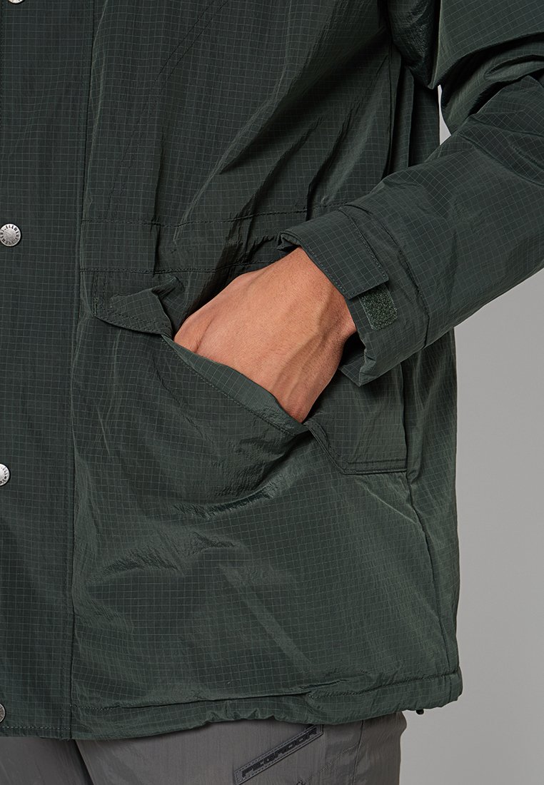 Ręka w kieszeni kurtki w ciemnozieloną kratę, kurtka ma napy i rzepy przy mankietach, osoba nosi szare spodnie.